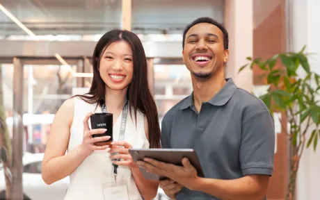 Two smiling colleagues standing side by side with a phone and tablet at work